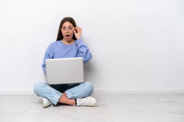 Young caucasian woman with laptop sitting on the floor isolated on white background with glasses and surprised