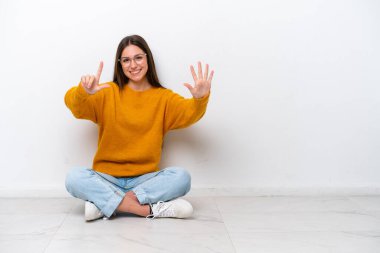 Yerde oturan genç kız, beyaz arka planda izole edilmiş, yediyi parmaklarıyla sayıyor.