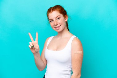 Mavi arka planda yara bandı takan genç, kırmızı bir kadın gülümsüyor ve zafer işareti gösteriyor.