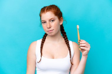 Young caucasian reddish woman brushing teeth isolated on blue background with sad expression