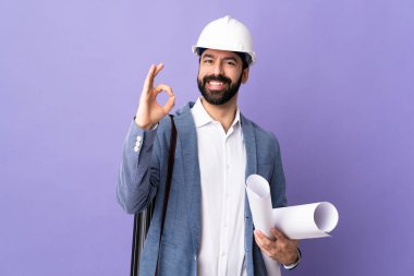 Miğferli genç mimar, elinde planlar, izole edilmiş mor arkaplan, parmaklarıyla tamam işareti gösteriyor.