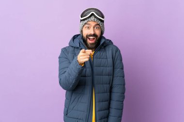 İzole edilmiş mor arka planda snowboard gözlüklü kayakçı şaşırmış ve ön tarafı işaret ediyor.
