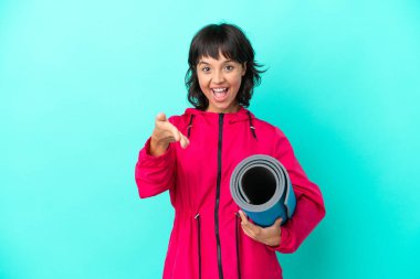 Genç kız yoga derslerine giderken mavi arka planda kapalı bir minder tutuyor ve iyi bir anlaşma için el sıkışıyor.