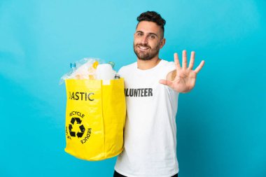 Mavi arka planda izole edilmiş plastik şişelerle dolu bir çanta taşıyan beyaz bir adam, parmaklarıyla beşi sayıyor.