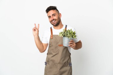 Bahçıvan elinde bir bitki tutuyor beyaz arka planda gülümsüyor ve zafer işareti gösteriyor.