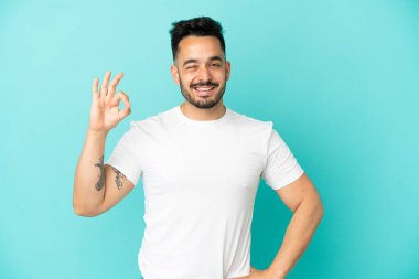 Mavi arka planda izole edilmiş genç beyaz adam parmaklarıyla tamam işareti gösteriyor.