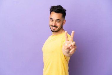 Mor arka planda izole edilmiş genç beyaz adam gülümsüyor ve zafer işareti gösteriyor.