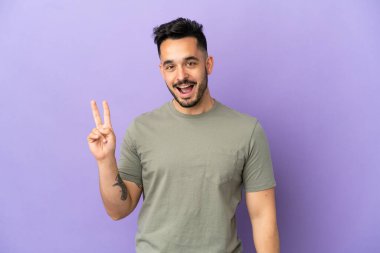 Mor arka planda izole edilmiş genç beyaz adam gülümsüyor ve zafer işareti gösteriyor.
