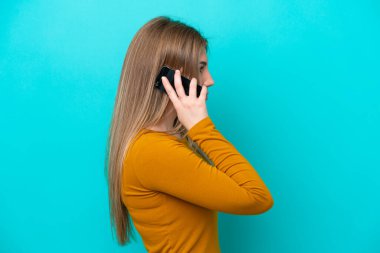 Mavi arka planda izole edilmiş genç beyaz bir kadın cep telefonuyla sohbet ediyor.