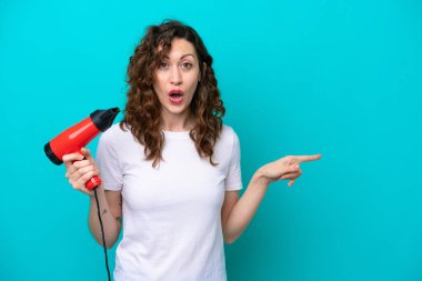 Mavi arka planda saç kurutma makinesi tutan beyaz bir kadın şaşırmış ve işaret ediyor.