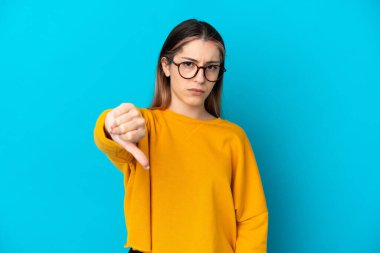 Mavi arka planda izole edilmiş genç beyaz kadın olumsuz ifadeyle baş parmağını aşağı gösteriyor.