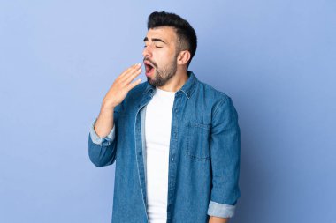 İzole edilmiş mavi arka planda beyaz bir adam esniyor ve ağzını eliyle kapatıyor.