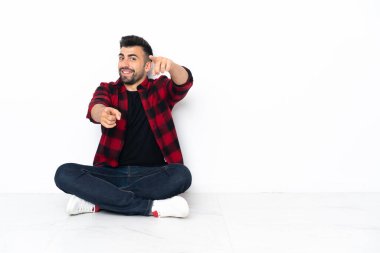 Young handsome man sitting on the floor points finger at you while smiling