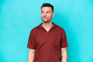 Kafkasyalı yakışıklı genç adam mavi arka planda izole edilmiş, yukarı bakarken şüpheleri var.