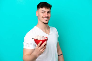 Mavi arka planda bir kase mısır gevreği tutan genç beyaz bir adam sırıtıyor.