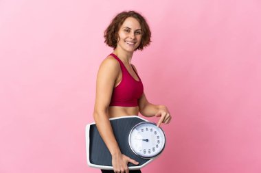 Pembe arka planda izole edilmiş genç bir İngiliz kadın elinde bir tartı makinesi tutuyor ve ona doğrultuyor.