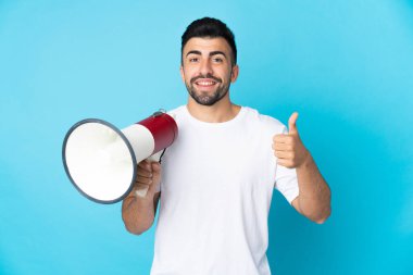 İzole edilmiş mavi arka planı olan beyaz bir adam elinde baş parmağıyla bir megafon tutuyor.