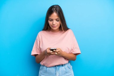 Mavi arka planda izole edilmiş genç beyaz kadın cep telefonuyla bir mesaj gönderiyor.