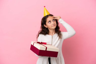 Küçük beyaz kız pembe arka planda izole edilmiş bir hediye tutuyor. Şüpheleri var ve kafası karışmış bir yüz ifadesi var.