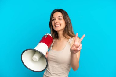 Mavi arka planda izole edilmiş genç beyaz kadın elinde bir megafonla gülümsüyor ve zafer işareti gösteriyor.