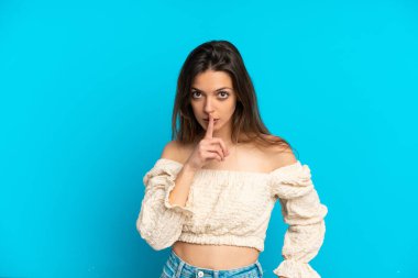 Mavi arka planda izole edilmiş beyaz bir kadın sessiz bir jest işareti gösteriyor. Parmağını ağzına sokuyor.