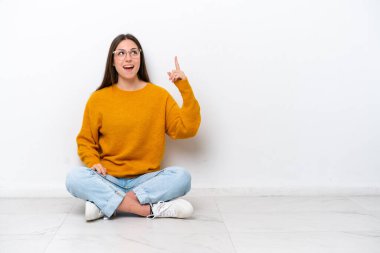 Genç kız beyaz arka planda izole bir şekilde yerde oturuyor. Yukarı bakıyor ve şaşırıyor.