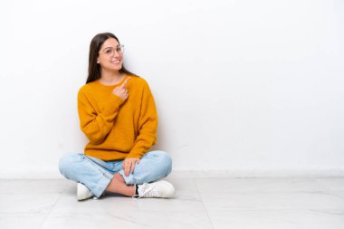 Beyaz arka planda izole bir şekilde yerde oturan genç kız bir ürünü sunmak için kenarı işaret ediyor.