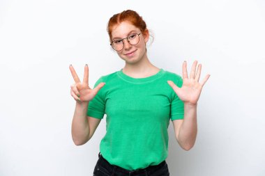 Beyaz arka planda izole edilmiş genç kızılımsı kadın parmaklarıyla sekiz sayıyor.