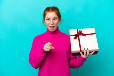 Kafkasyalı genç bir kadın elinde mavi arka planda izole edilmiş bir hediye tutuyor şaşırmış ve ön tarafı işaret ediyor.