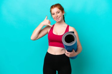 Genç spor kırmızısı bir kadın yoga derslerine giderken elinde mavi arka planda izole edilmiş bir minder telefon jesti yapıyor. Bana geri dönüş işareti ver.