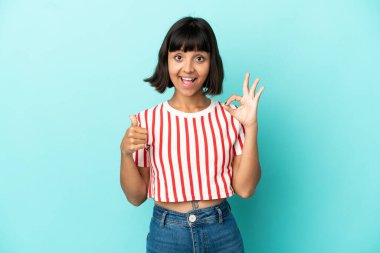Mavi arka planda izole edilmiş genç melez kadın işaret ve baş parmak işareti gösteriyor.