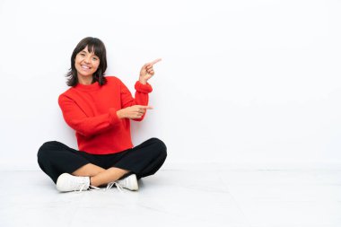 Genç melez bir kadın, beyaz arka planda izole bir şekilde yerde oturuyor. Parmaklarını kenarı gösteriyor ve bir ürün sunuyor.