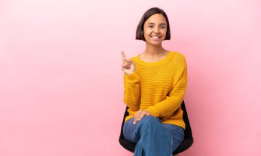 Genç melez kadın pembe arka planda izole edilmiş bir sandalyede oturmuş harika bir fikre işaret ediyor.