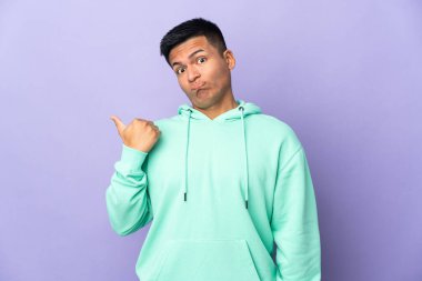 Genç Ekvadorlu adam, mor arka planda izole edilmiş mutsuz ve kenarı işaret ediyor.