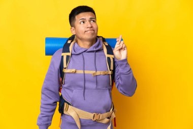 Young mountaineer man with a big backpack isolated on yellow background with fingers crossing and wishing the best