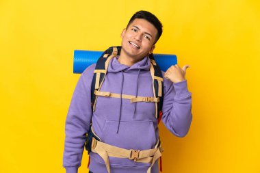 Sarı arka planda izole edilmiş büyük sırt çantalı genç dağcı bir ürünü sunmak için kenarı işaret ediyor.