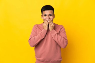 Genç Ekvadorlu adam sarı arka planda izole edilmiş mutlu ve gülümseyen elleriyle ağzını kapatıyor.