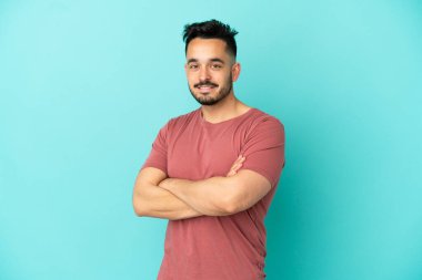 Genç beyaz adam mavi arka planda izole edilmiş, kolları bağlı ve ileriye bakıyor.