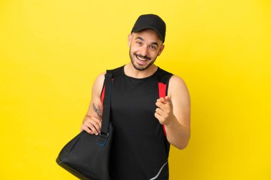 Spor çantalı genç beyaz adam sarı arka planda izole edilmiş şaşırmış ve ön tarafı işaret ediyor.