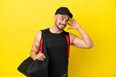Spor çantalı genç beyaz adam sarı arka planda izole edilmiş bir şekilde elini kulağına koyarak bir şeyler dinliyor.