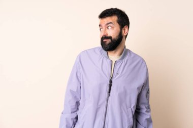 Sakallı beyaz bir adam, soyutlanmış arka planda ceket giyiyor. Şüpheli hareketler yapıyor.