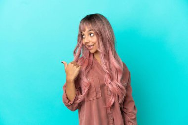 Mavi arka planda izole edilmiş pembe saçlı melez bir kadın bir ürünü sunmak için kenarı işaret ediyor.