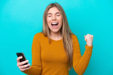 Mavi arka planda izole edilmiş genç beyaz kadın cep telefonu kullanıyor ve zafer jesti yapıyor.
