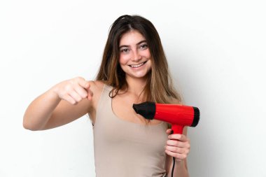 Beyaz arka planda izole edilmiş saç kurutma makinesini tutan genç beyaz kadın kendine güvenen bir ifadeyle seni işaret ediyor.
