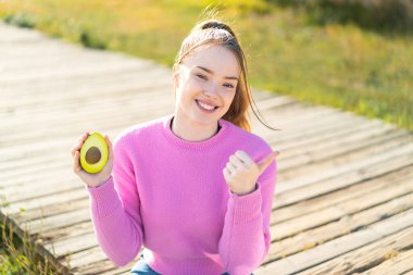 Dışarıda elinde avokado tutan genç ve güzel bir kız bir ürün sunmak için yan tarafı işaret ediyor.