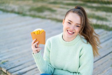 Genç ve güzel bir kız elinde patates kızartması ile dışarıda gülümsüyor.