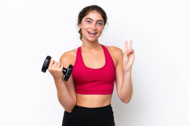 Beyaz arka planda halter kaldıran genç beyaz kadın gülümsüyor ve zafer işareti gösteriyor.