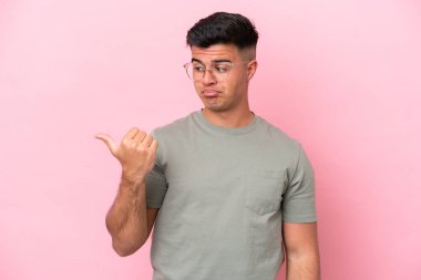 Pembe arka planda izole edilmiş genç beyaz yakışıklı adam mutsuz ve bir tarafı işaret ediyor.