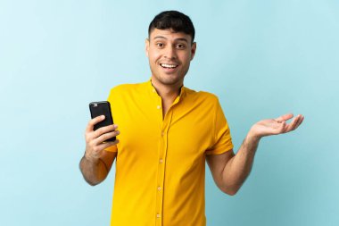 Cep telefonu kullanan genç Kolombiyalı adam şok olmuş yüz ifadesine sahip mavi arka planda izole edilmiş.