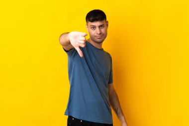 Sarı arka planda izole edilmiş genç Kolombiyalı adam olumsuz ifadelerle baş parmağını aşağı gösteriyor.